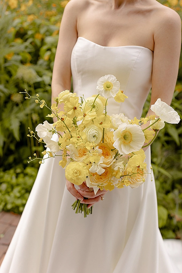 Bridal bouquet of yellow and white flowers with poppies, ranunculus, roses and orchids, held against a strapless dress in garden greenery