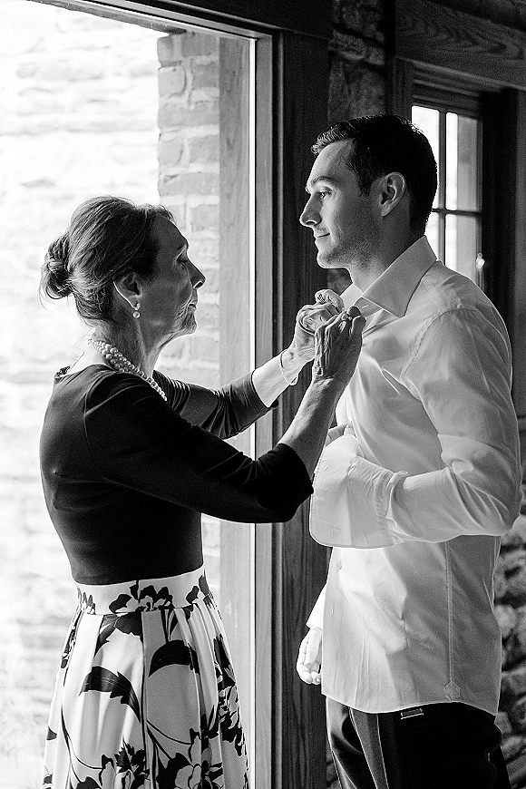 Groom getting ready as his mother helps button his dress shirt, cufflinks catching window light in a rustic room with stone wall