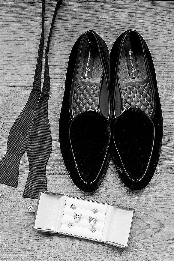 Groom accessories flat lay with black dress shoes, bow tie, cufflinks, and stud set arranged neatly on a wood floor