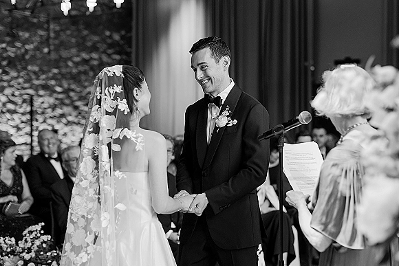 Wedding vows as bride in a lace veil and groom in tux hold hands, reading printed sheets into a microphone before guests indoors