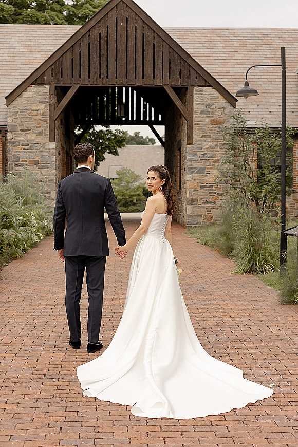 Couple portrait of bride and groom holding hands walking away on a brick walkway by a stone building, her strapless gown train flowing