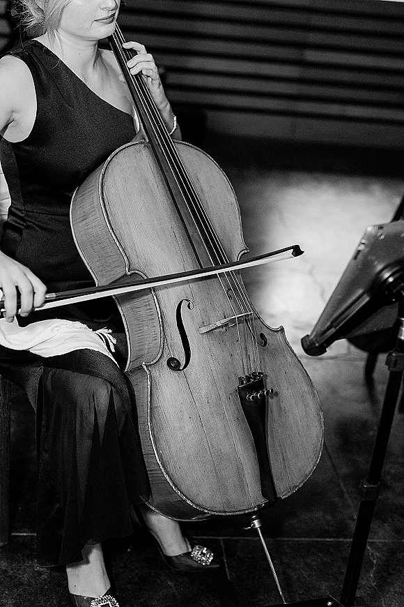 Wedding musician plays cello with bow beside a music stand, wearing a formal black dress on indoor venue steps