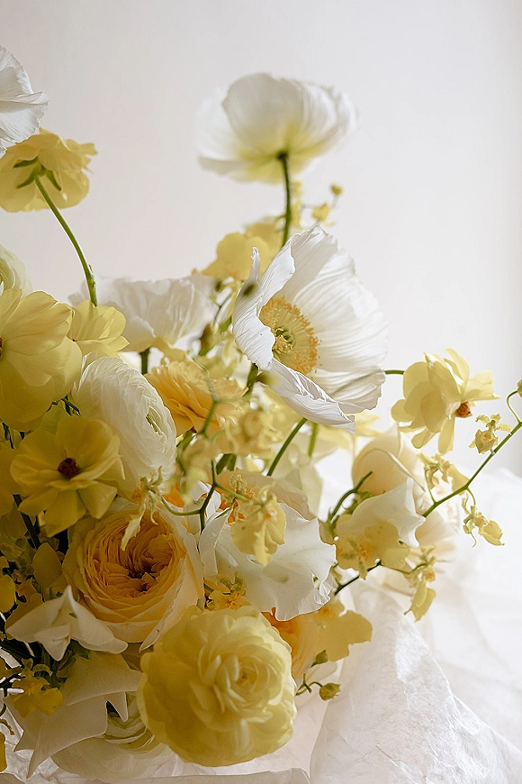Wedding bouquet with yellow and white bouquet tones, featuring yellow roses and white poppies, resting on white linen fabric backdrop