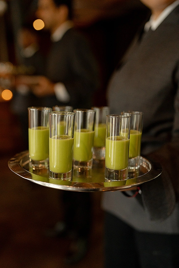 Wedding cocktail tray of green shooters and shot glasses on a metal serving tray, held by suited staff in a warm-lit indoor reception