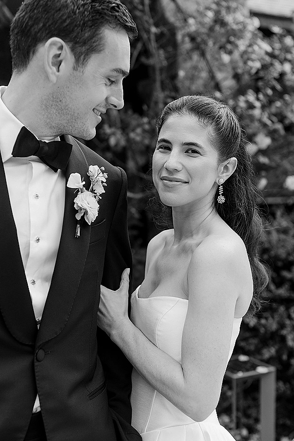 Wedding couple portrait in black and white, bride in a strapless dress embracing groom in tuxedo with boutonniere against blurred greenery