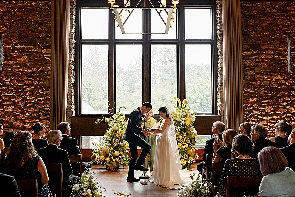 Wedding vows as bride in veil and groom in black tux hold hands under floral arch with yellow blooms in a stone-walled venue