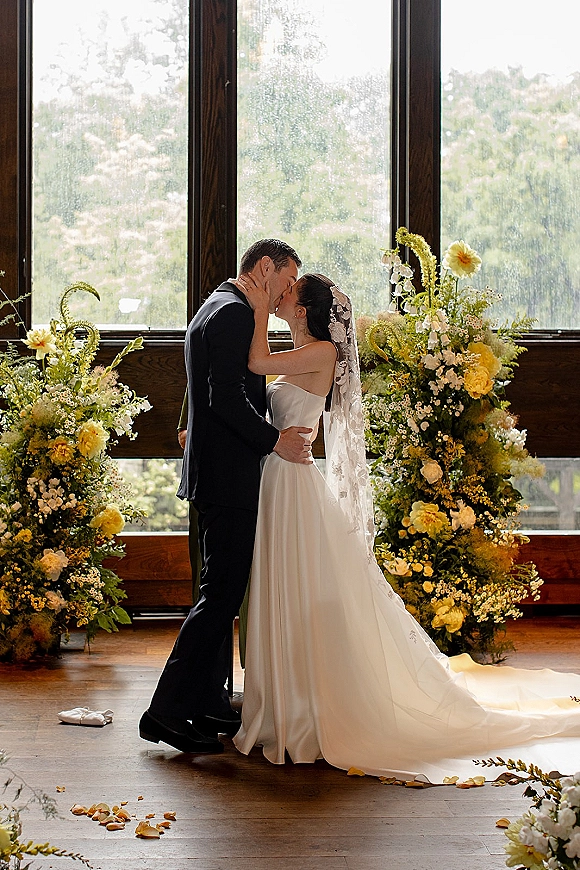 Wedding kiss portrait of the bride and groom kissing in window light, her veil softly backlit, framed by yellow and white florals indoors