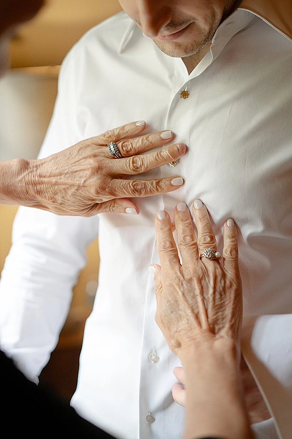 Groom getting ready as manicured hands fasten buttons on his white dress shirt with a collar pin in a warm, neutral indoor room