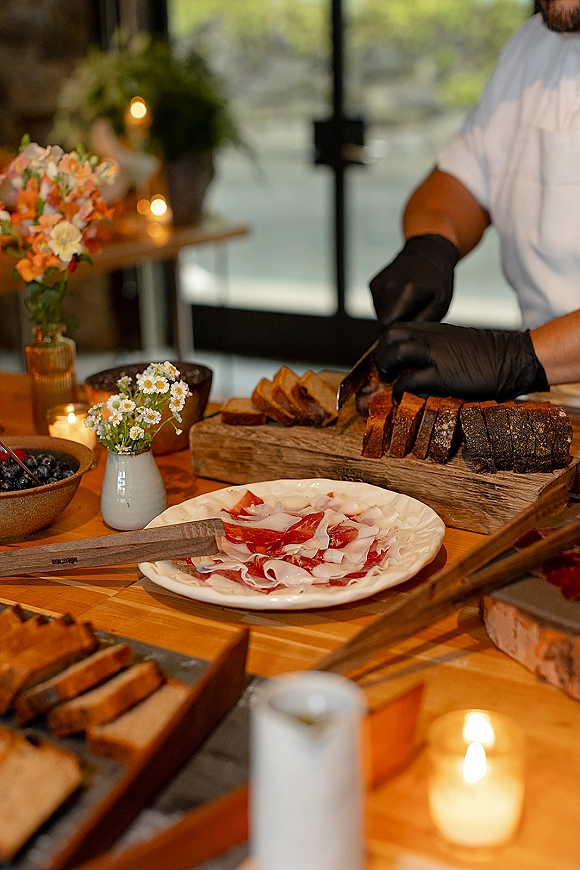 Wedding catering station with live carving station bread and charcuterie on wooden boards, candles and bud vases by glass doors with greenery
