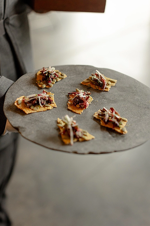 Wedding appetizers arranged as passed hors d'oeuvres on crackers with shredded topping, served on a gray platter held by a suit jacketed server