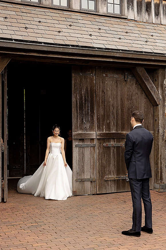 Wedding first look as bride walking to groom in a strapless dress with long train, beside rustic barn doors in a brick courtyard