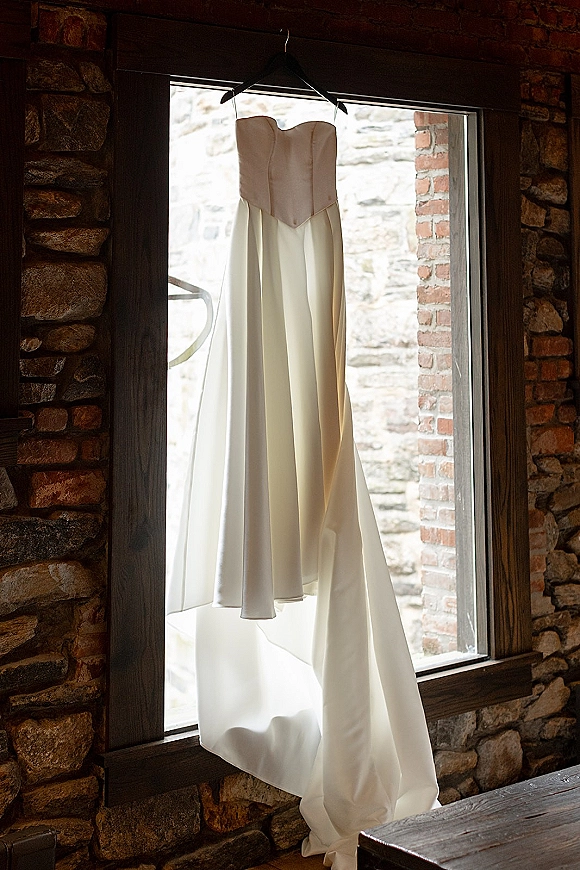 Wedding dress hanging in a window, strapless satin gown with a corset bodice on a hanger against stone and brick walls in natural light