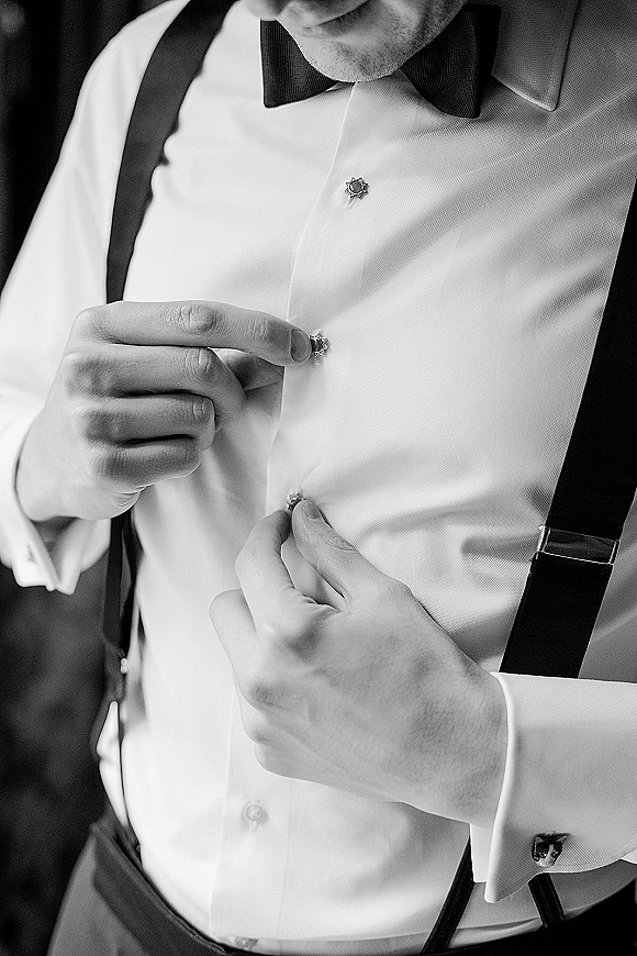 Groom getting ready, buttoning his dress shirt with shirt studs, cufflinks, and a black bow tie with suspenders in a dark room