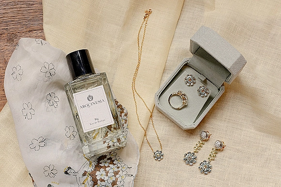 Wedding jewelry flatlay featuring an engagement ring, diamond earrings and gold necklace on beige linen with perfume bottle and jewelry box on wood surface