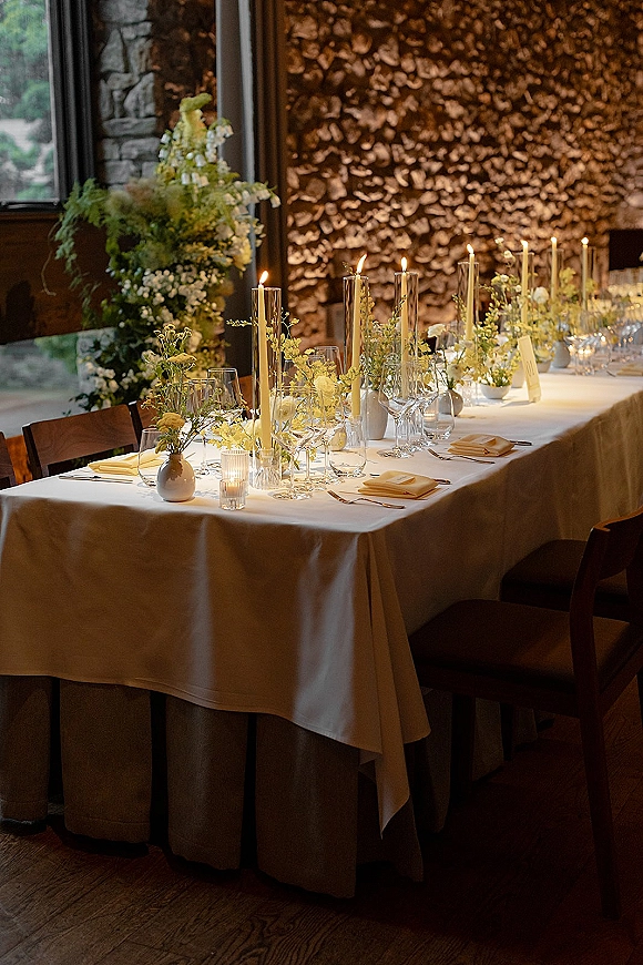 Reception tablescape with long head table decor, ivory taper and votive candles, yellow flowers in bud vases by a stone wall window