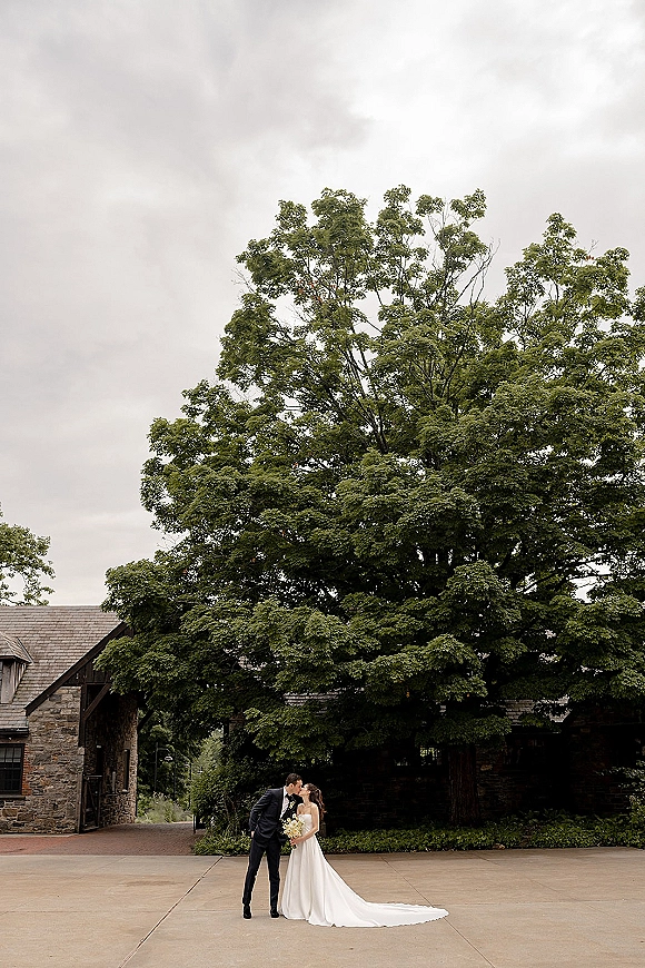 Wedding kiss portrait of bride and groom kissing, her bouquet and dress train flowing beside a stone courtyard with large tree backdrop