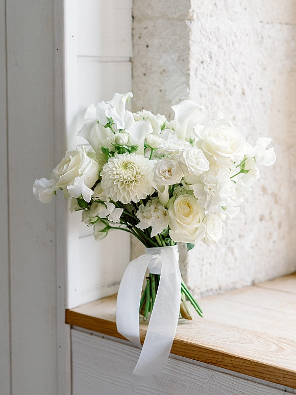 White bridal bouquet of roses, dahlia and sweet peas with a ribbon bow on a windowsill in soft window light against a stone wall