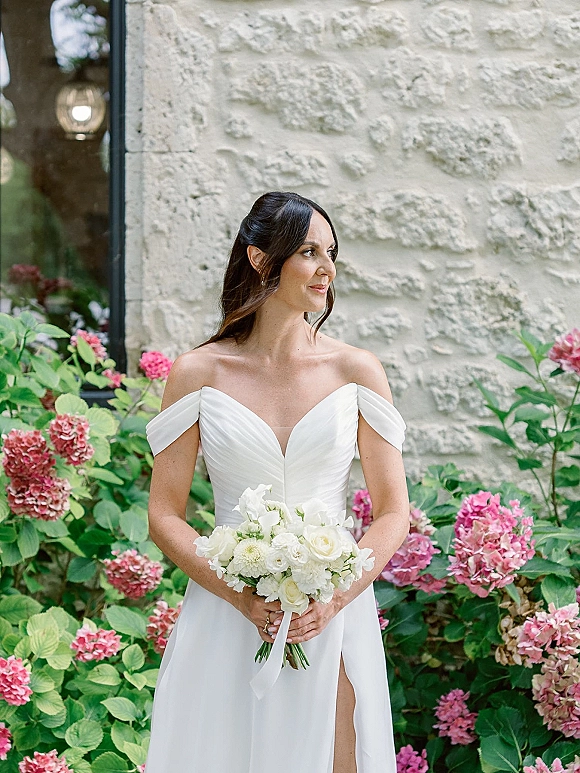 Bridal portrait of a bride in an off the shoulder wedding dress holding a white rose bouquet with ribbon near pink hydrangeas and a stone wall window