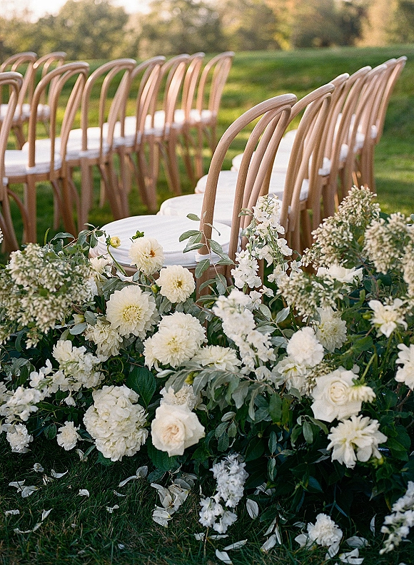 Ceremony aisle decor with outdoor ceremony chairs, wood chairs and white cushions, lined with white rose and eucalyptus florals on grass lawn