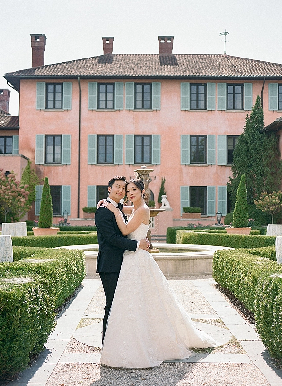 Couple portrait of bride and groom hugging by a garden fountain, her veil over a strapless gown as he wears a black tuxedo near a villa path