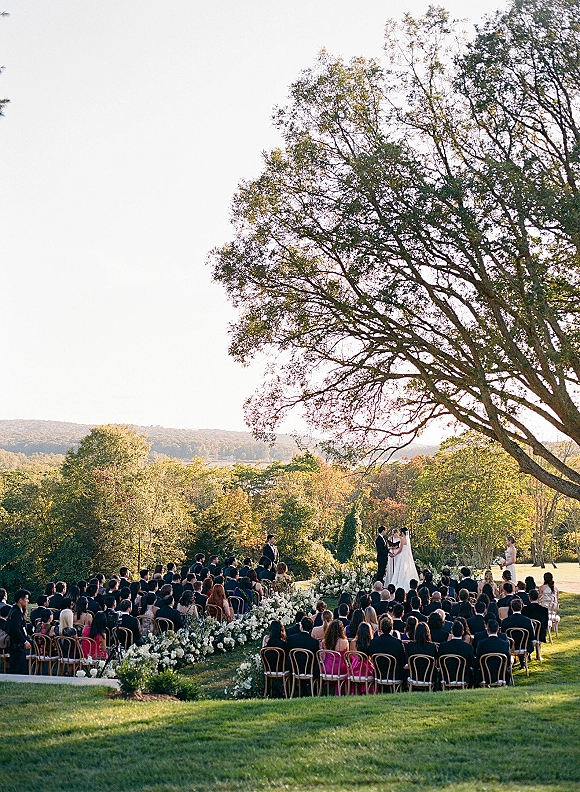 Outdoor wedding ceremony with bride in veil and groom in tux at the altar, white aisle flowers lining chairs on a green lawn under a large tree