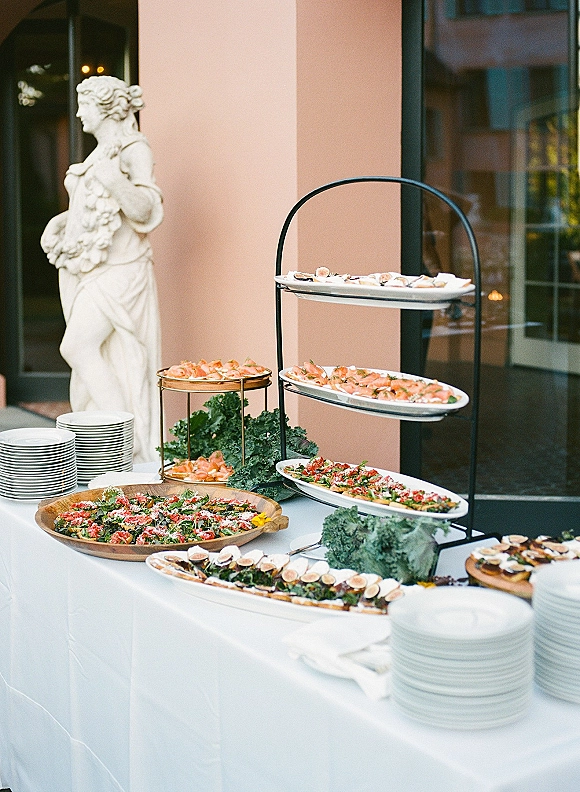 Wedding cocktail hour wedding appetizer display on a white tablecloth with tiered stand, platters, stacked plates, and kale garnish on a patio near columns
