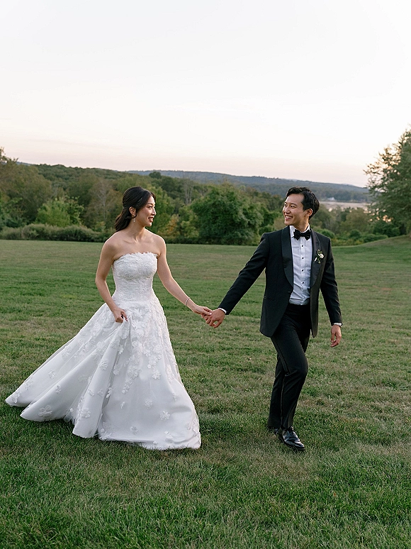 Couple portrait of bride and groom holding hands while walking in a grassy field, her strapless lace gown and his tuxedo at sunset