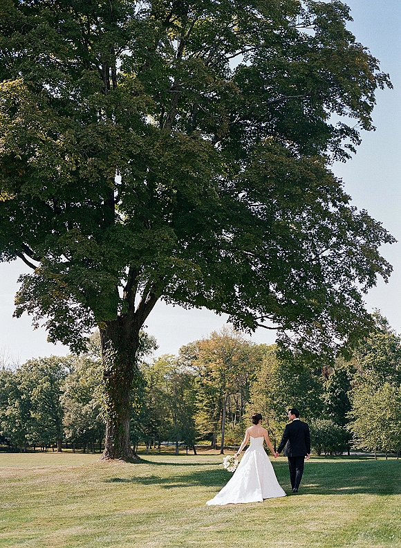 Couple portrait of bride and groom walking away holding hands, her strapless dress train and white bouquet under a large park tree