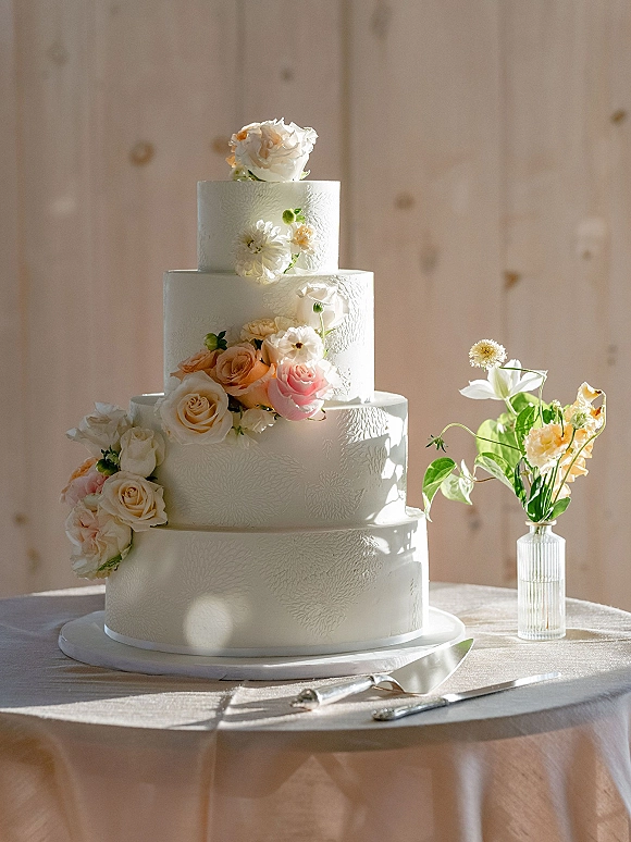 Wedding cake with textured buttercream, tiered white design and blush roses, on a stand with knife and server against sunlit wood wall
