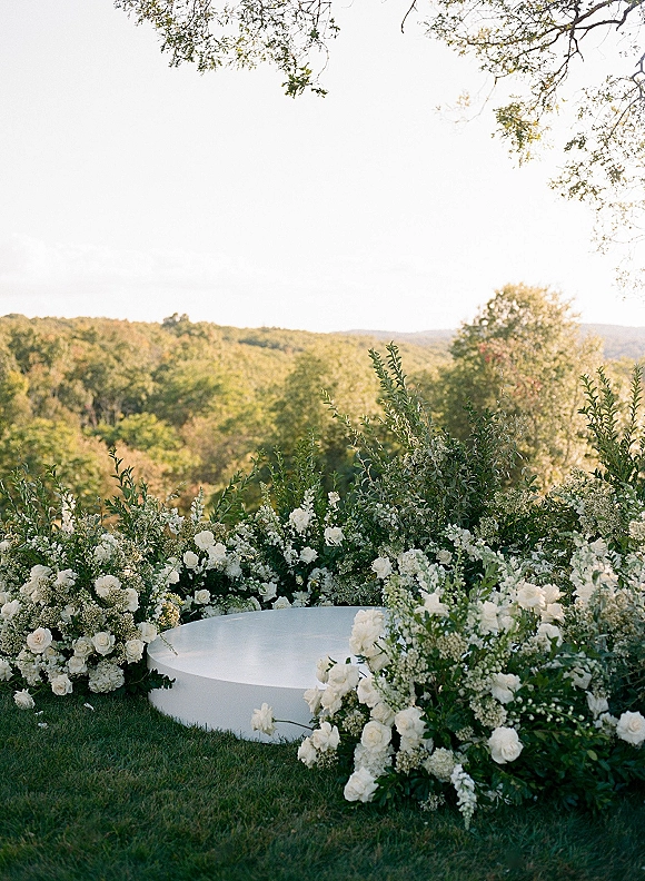 Wedding ceremony backdrop with white floral arrangements and greenery garland on a round white platform stage on a grassy lawn with forested hills