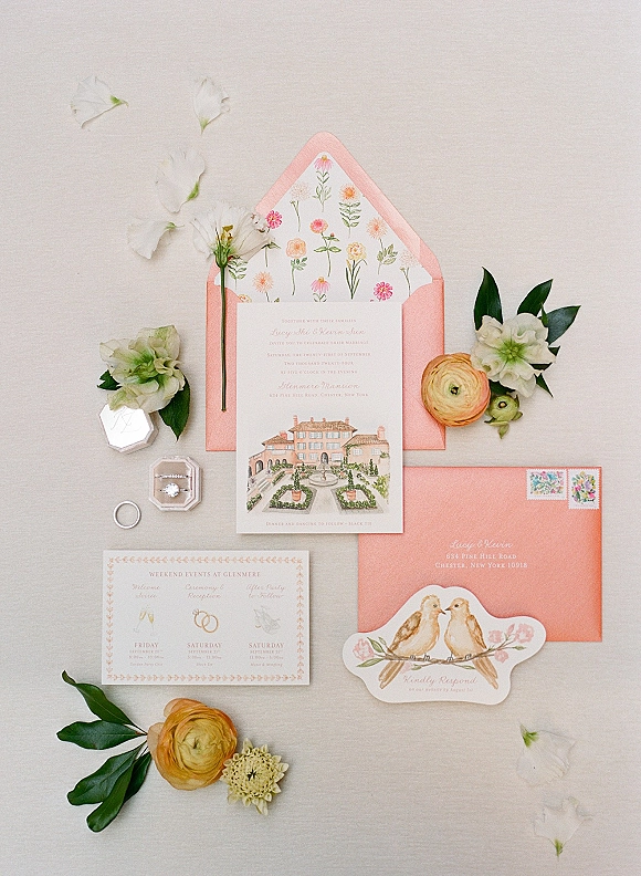 Invitation suite flat lay with peach envelopes, floral liners, RSVP and details cards, wax seal, rings, and ranunculus on light fabric backdrop