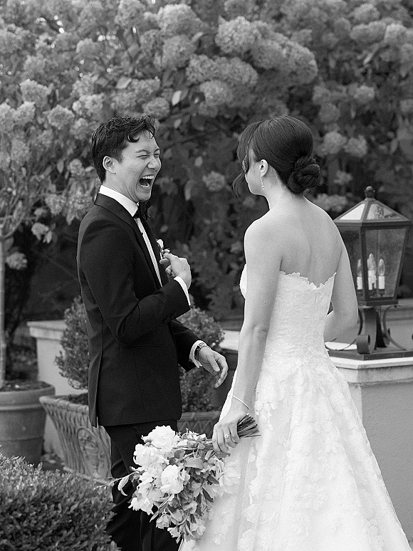 First look moment as bride in a strapless lace gown holds a bouquet while groom in tux with boutonniere laughs by garden shrubs