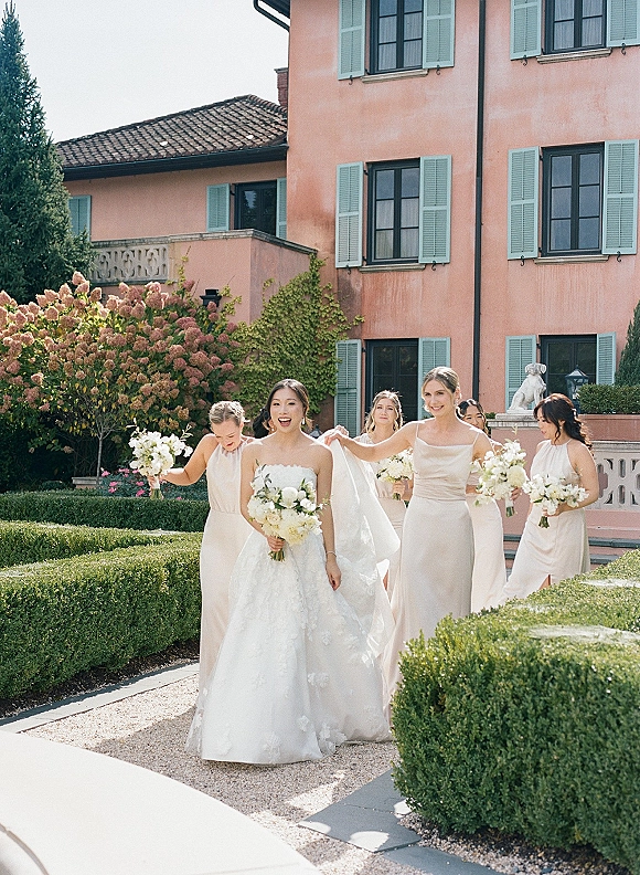 Bride with bridesmaids walking as a bridal party while one adjusts her long veil, bouquets in hand along a gravel garden path by a villa