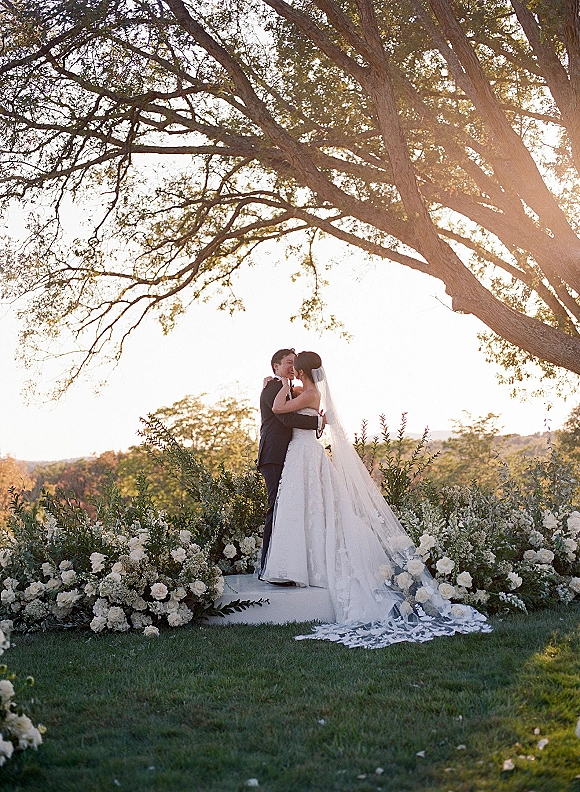 Wedding kiss as bride in wedding dress and cathedral veil embraces groom in tuxedo beneath a large tree at sunset on a lawn