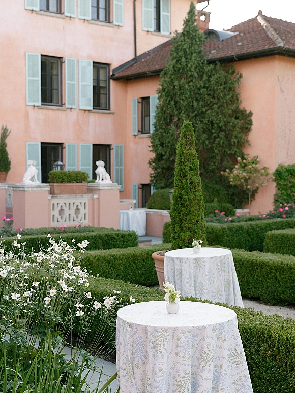 Cocktail hour setup with outdoor cocktail tables in a villa courtyard, featuring patterned linens and bud vases beside manicured hedges and blue shutters