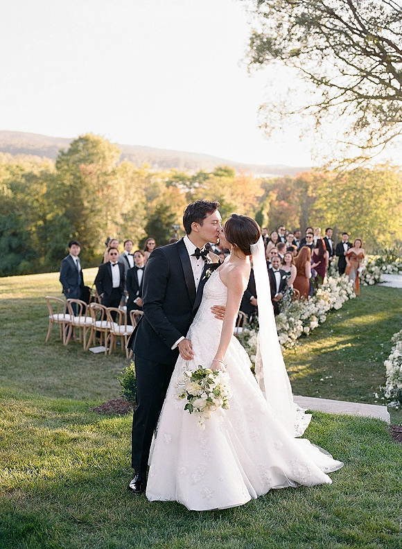 Wedding kiss as bride in a strapless dress and veil holds a white bouquet, kissing groom in tuxedo on an aisle with guests and hills behind