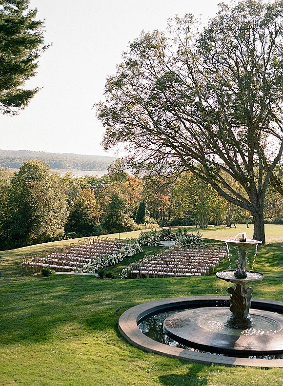 Outdoor ceremony setup with curved ceremony chairs and white aisle flowers on a lawn beside a fountain, under a large tree with hills beyond