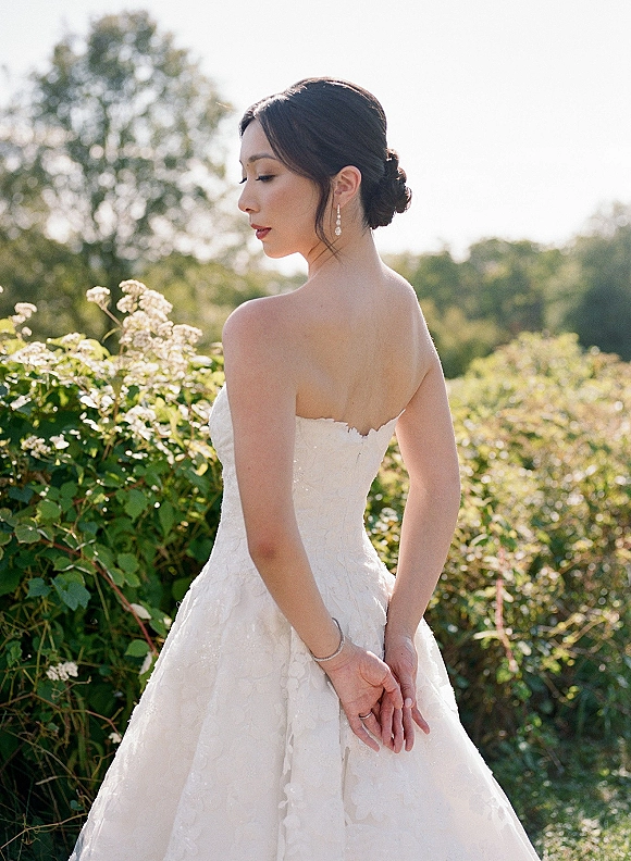 Bridal portrait of a bride in a strapless wedding dress and lace ball gown, looking over her shoulder in a sunlit garden setting