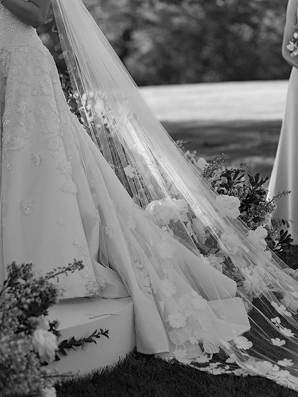 Bridal veil with floral applique veil draped over a rose-and-greenery bouquet, lace dress trailing on garden lawn by stone steps