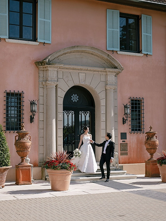 Couple portrait of bride and groom holding hands, bride with white bouquet in strapless gown and groom in tux at villa entrance steps.