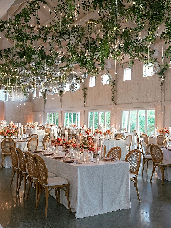 Reception tablescape with wedding reception table decor, white tablecloths, floral centerpieces and floating candles under hanging greenery in a white barn