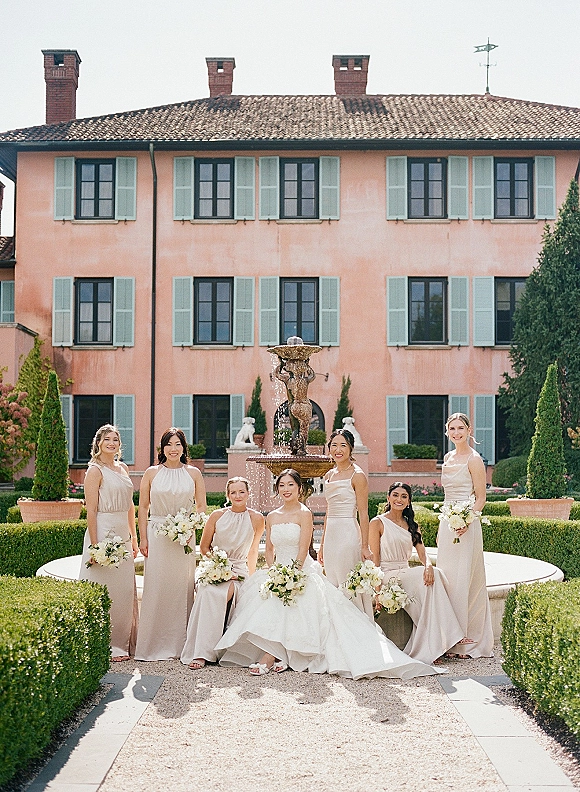 Bridesmaid group photo with bride in strapless gown and bridesmaids in champagne dresses holding white bouquets by a villa fountain