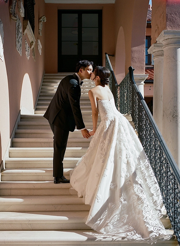 Wedding kiss portrait of bride and groom kissing, holding hands on a sunlit staircase with wrought iron railing and arched doorway backdrop