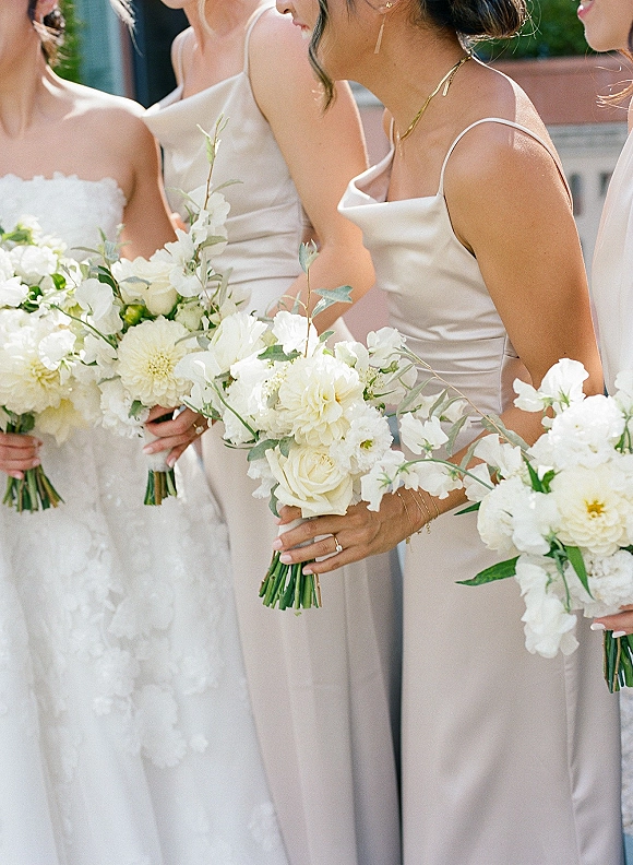 Bridesmaid bouquets of white flowers and greenery held by bridesmaids in champagne dresses beside the bride on a sunlit terrace