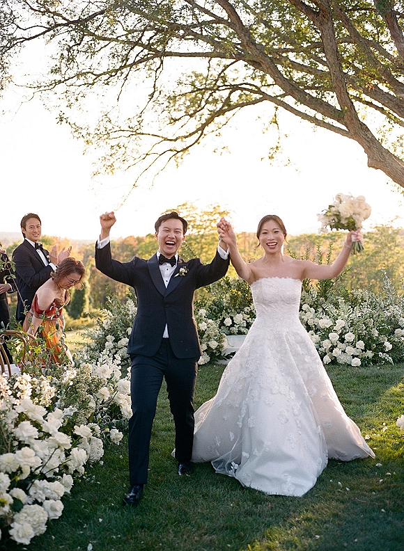 Wedding recessional as bride and groom walk hand in hand down a garden aisle at sunset, bouquet raised, guests cheering under trees