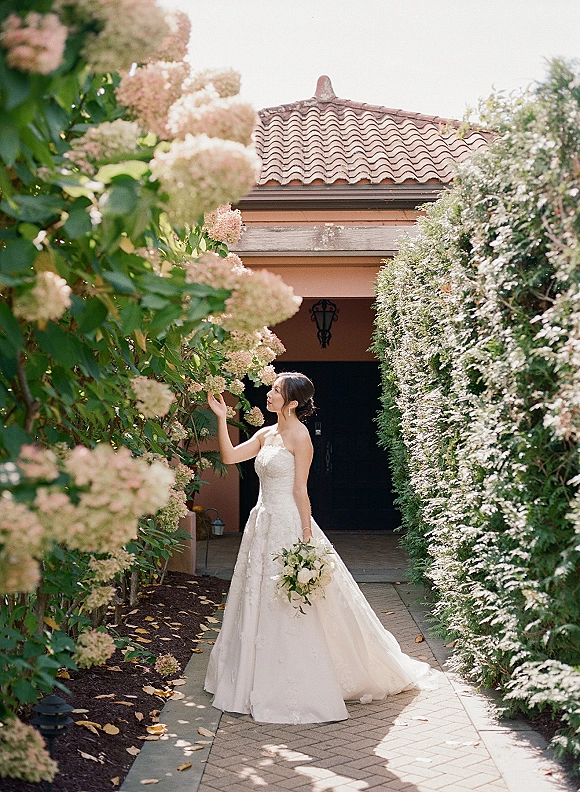 Bridal portrait of a bride holding bouquet, in a strapless lace wedding dress with drop earrings on a sunlit garden walkway by a stucco villa