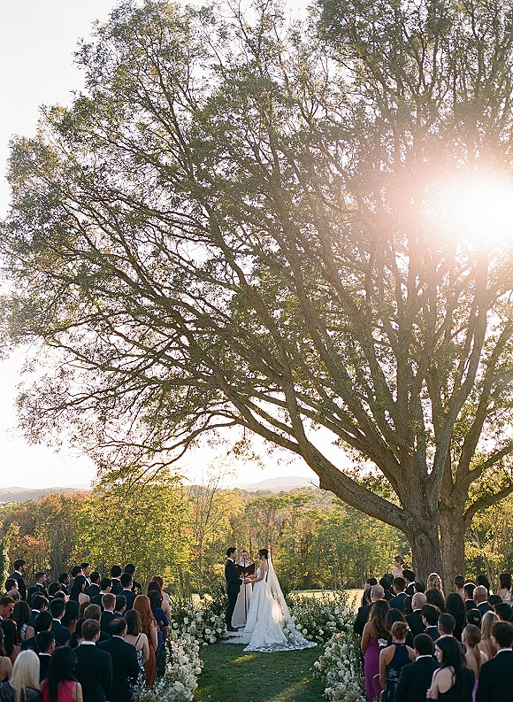 Wedding ceremony as bride and groom exchange vows beneath a large tree, bride’s veil flowing with a white floral arch in sunset light