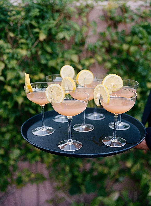 Wedding cocktails on a black serving tray in coupe glasses with lemon slice citrus garnish against a lush green ivy wall