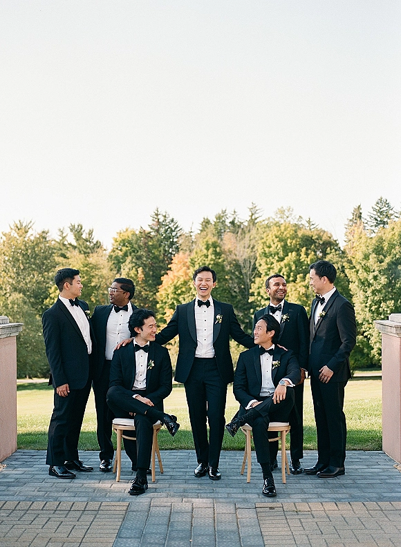 Groomsmen portrait with men in black tuxedos and bow ties, seated and standing on chairs on a stone patio with trees and columns behind