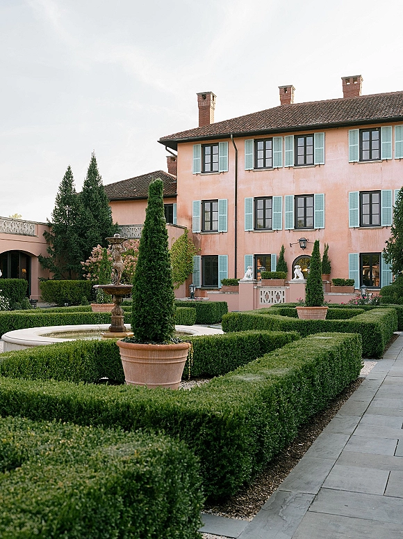 Villa courtyard with a stone fountain centered among trimmed boxwood hedges and terracotta planters against a stucco villa facade